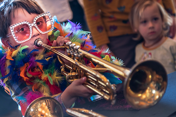 A child wearing wild sunglasses, and a feather boa plays the trumpet